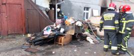 Strażacy w ciemnych mundurach i czerwonych hełmach stoją przed budynkiem przed którym zostały wyrzucone spalone i nadpalone materiały wyposażenia wnętrz: szafy, krzesła, półki. W tle budynek częściowo drewniany, częściowo murowany z szarą elewacją okopcony sadzą.