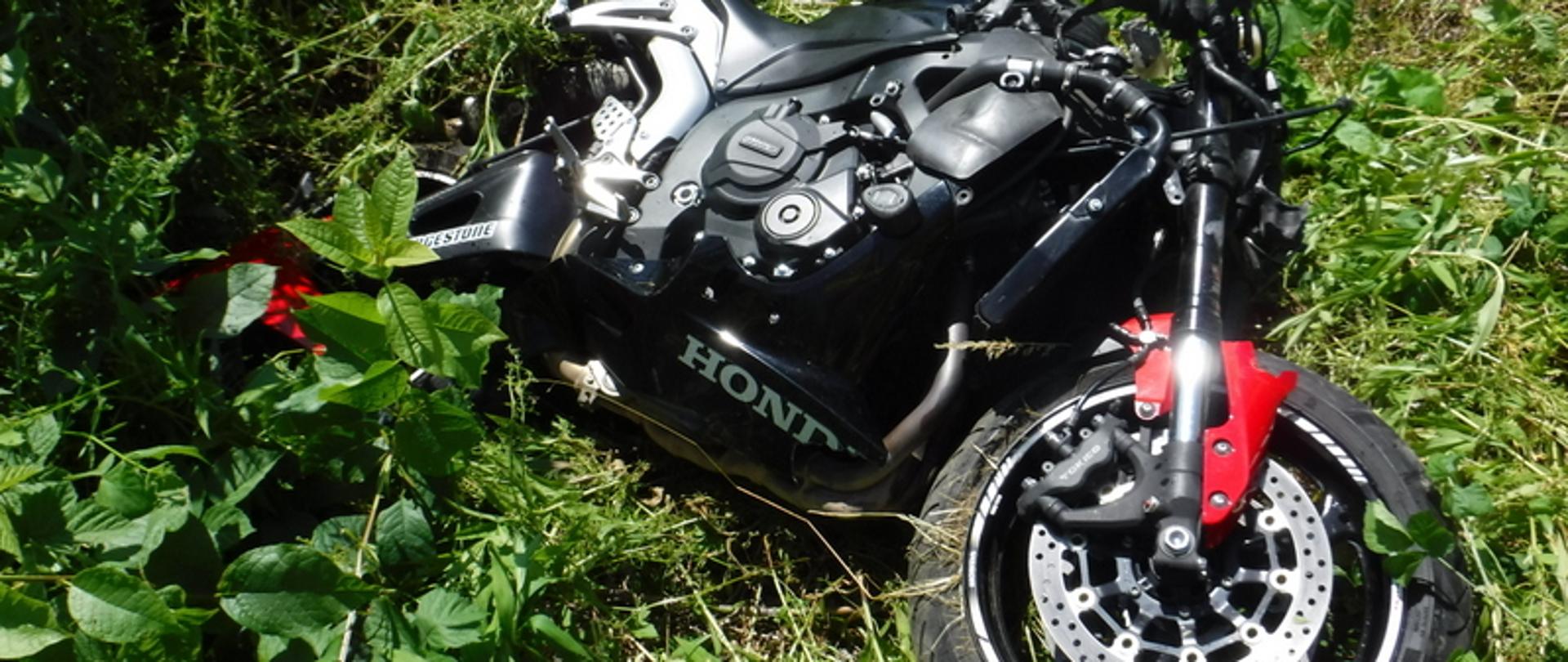 Zdjęcie przedstawia leżący motocykl marki Honda koloru czarnego z czerwonymi błotnikami. Motocykl leży na trawie na lewym boku. Widoczne zniszczenia bryły pojazdu z prawej strony - skrzywiona kierownica, uszkodzone siedzisko, uszkodzony błotnik.