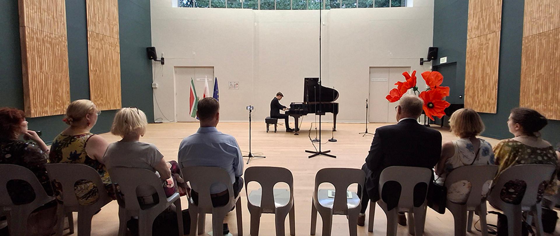 Concert, a man in black clothes plays piano, group of 7 guests sitting and listening to the music, two empty chairs in the middle of the row, 3 big red flowers to the right and three flags (SA, Poland and EU) to the left