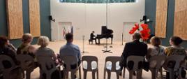 Concert, a man in black clothes plays piano, group of 7 guests sitting and listening to the music, two empty chairs in the middle of the row, 3 big red flowers to the right and three flags (SA, Poland and EU) to the left