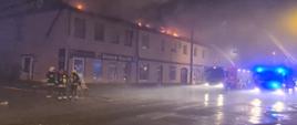 Zdjęcie przedstawia widok na palące się dachy i poddasza budynków. Z lewej strony widzimy trzech strażaków prowadzących działania ratowniczo - gaśnicze, natomiast z prawej strony widać samochody ratowniczo - gaśnicze.