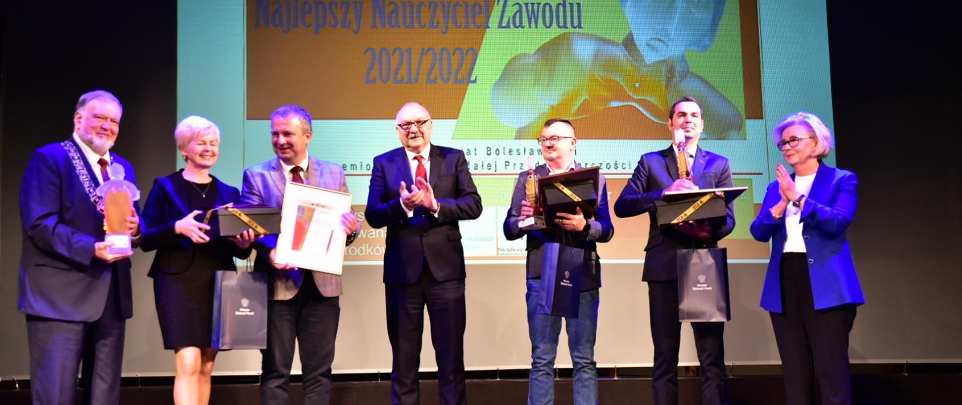 Grupa kilku osób trzymających książki stoi pod ścianą, na której jest napis Najlepszy Nauczyciel Zawodu Powiatu Bolesławieckiego.