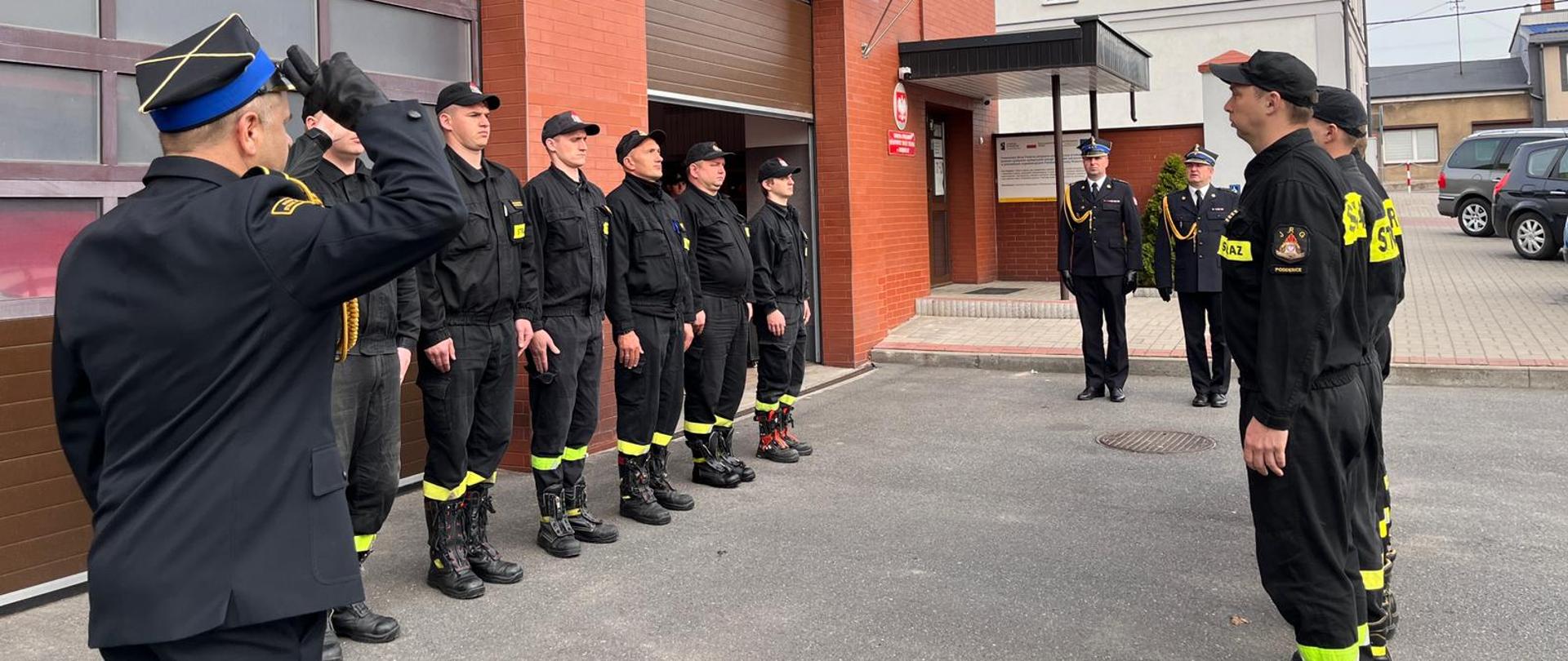 Przed budynkiem komendy w dwóch rzędach naprzeciwko siebie stoją strażacy w czarnych strojach strażackich