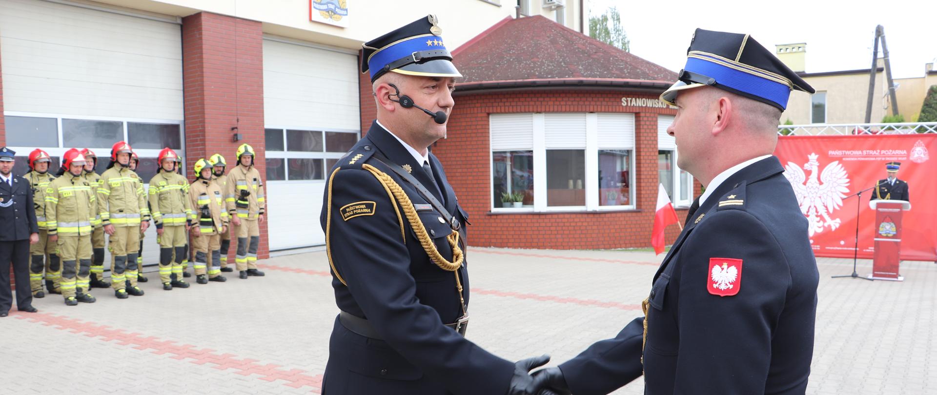 strażak ściska rękę drugiemu strażakowi, w tle komenda i grupa strażaków w mundurach bojowych i hełmach