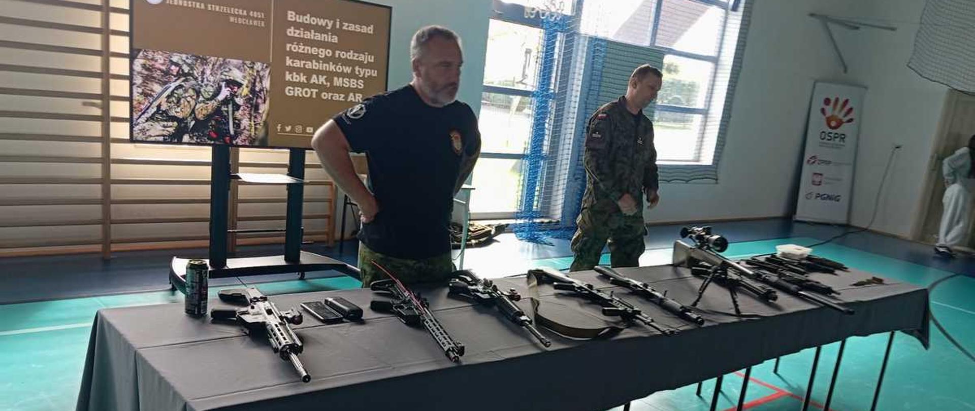 Zdjęcie przedstawia wystawę broni palnej zorganizowaną w hali sportowej, przygotowaną prawdopodobnie na potrzeby pokazu lub szkolenia dla młodzieży [image_nKurWG.png]. Kadr obejmuje długi stół z prezentowanymi egzemplarzami oraz dwóch prowadzących w tle [image_nKurWG.png].Oto szczegółowy opis poszczególnych elementów zdjęcia:Stół z bronią (pierwszy plan): W poprzek sali ustawiony jest długi stół przykryty ciemnoszarym obrusem [image_nKurWG.png]. Równo, w jednym rzędzie, ułożonych jest na nim około ośmiu sztuk długiej broni palnej (karabinki automatyczne, karabiny wyborowe z lunetami oraz repliki) [image_nKurWG.png]. Wszystkie lufy skierowane są w prawą stronę [image_nKurWG.png]. Na stole, obok broni, leżą także zapasowe magazynki, małe akcesoria w plastikowym pudełku oraz puszka z napojem po lewej stronie [image_nKurWG.png].Osoby w tle: Za stołem stoją dwaj mężczyźni, którzy nadzorują wystawę [image_nKurWG.png]:Po lewej stoi postawny, brodaty mężczyzna w czarnym t-shircie i spodniach w kamuflażu [image_nKurWG.png]. Trzyma ręce oparte na biodrach i patrzy w stronę prawej krawędzi stołu [image_nKurWG.png].Po prawej, nieco głębiej w sali, stoi żołnierz w pełnym umundurowaniu polowym (kamuflaż pantera) z polską flagą na ramieniu [image_nKurWG.png]. Idzie powoli wzdłuż stołu [image_nKurWG.png].Ekran multimedialny: Za plecami mężczyzn, po lewej stronie, stoi duży monitor na stojaku [image_nKurWG.png]. Wyświetla on planszę informacyjną z logo „Jednostka Strzelecka 4051 Włocławek” [image_nKurWG.png]. Tekst na ekranie brzmi: „Budowy i zasad działania różnego rodzaju karabinków typu kbk AK, MSBS GROT oraz AR” [image_nKurWG.png]. Obok tekstu widoczne jest zdjęcie żołnierza w kamuflażu [image_nKurWG.png].Otoczenie: Prezentacja odbywa się na zielonym parkiecie sali gimnastycznej z wyznaczonymi czerwonymi liniami boiska [image_nKurWG.png]. W tle widać drewniane drabinki gimnastyczne na ścianie, duże okna dachowe wpuszczające jasne światło oraz zawieszoną wysoko tablicę do koszykówki z obręczą [image_nKurWG.png]. Po prawej stronie sali stoi biały baner z logo „OSPR” i „PGNiG” [image_nKurWG.png].