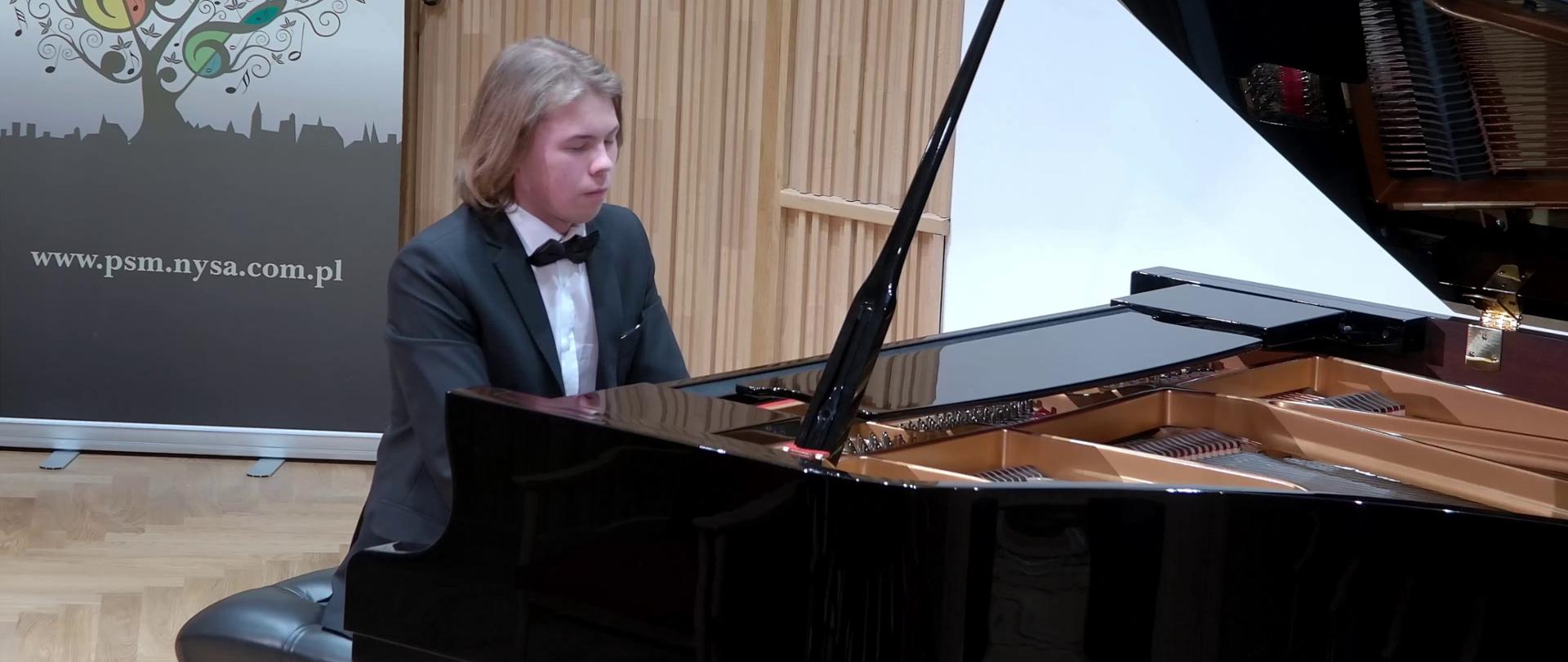 Zdjęcie. Młody pianista gra na fortepianie podczas koncertu na estradzie sali koncertowej. W tle baner PSM Nysa.