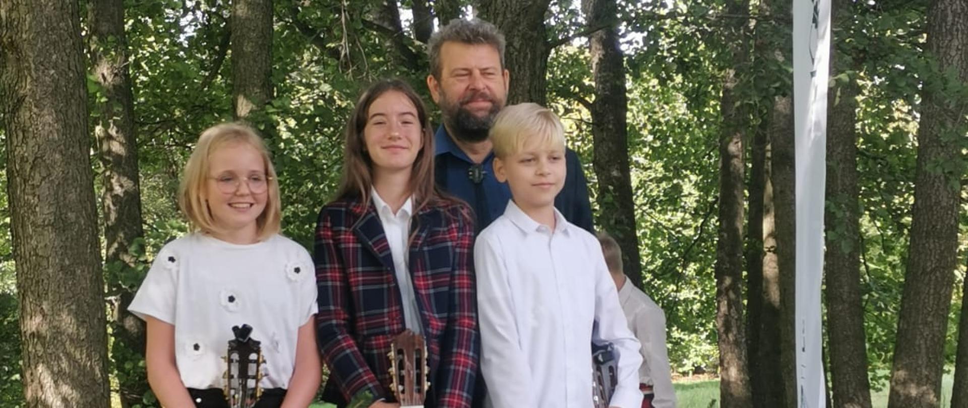 Uczniowie z klasy gitary Witolda Kozakowskiego (Natalia Skrzydło, Dobrawa Wers-Skawina i Cezary Petelski) stojący na tle drzew i trzymający gitary wraz z upominkami
