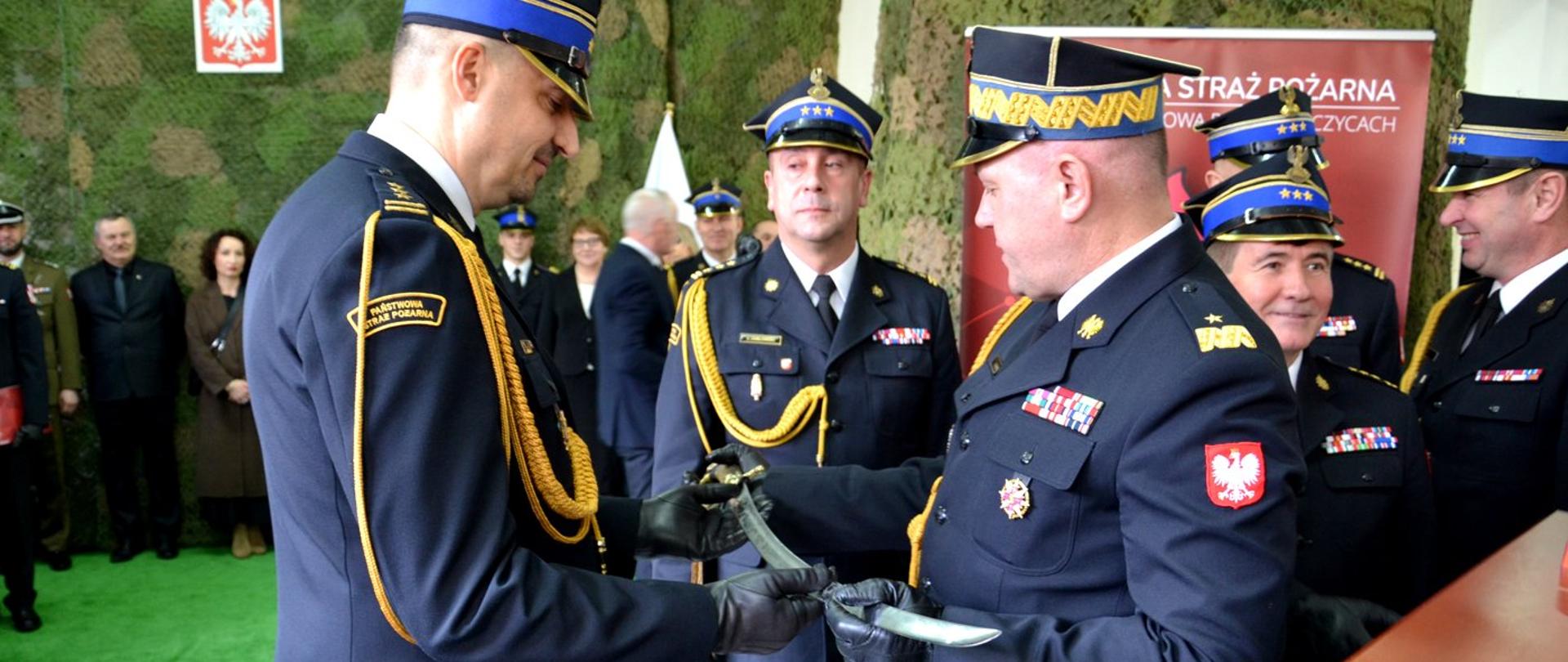Zdjęcie zrobione w pomieszczeniu, podczas uroczystości pożegnania odchodzącego na emeryturę komendanta Powiatowego Państwowej Straży Pożarnej. Na zdjęciu oficerowie Państwowej Straży Pożarnej, wśród nich generał, wręczają odchodzącemu komendantowi pamiątkową szablę. 