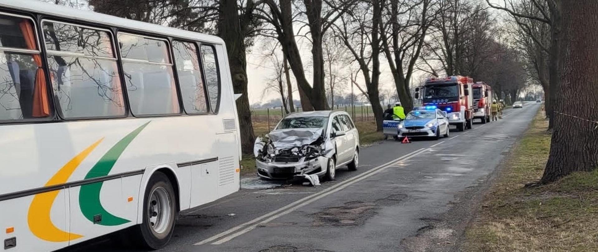 Zdjęcie na pierwszym planie przedstawia autobus oraz samochód osobowy po kolizji drogowej. Na drugim planie radiowóz policyjny, dwa wozy straży pożarnej oraz funkcjonariusze policji oraz strażacy podczas czynności.
