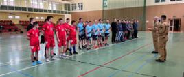 Drużyny uczestniczące w turnieju sportowym stoją w szeregu podczas ceremonii otwarcia, a przedstawiciel Państwowej Straży Pożarnej wygłasza przemówienie w hali sportowej.