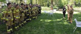 Ćwiczenia w Pakosławiu. Zbiórka końcowa, podczas której podsumowywany jest przebieg ćwiczeń. Na przeciwko ustawionych w dwuszeregu strażaków stoją funkcjonariusze KP PSP w Rawiczu i przedstawicielka zarządzającego obiektem. W tle park.