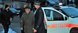 Widok z przodu. Na pierwszym planie strażak-emeryt stoi obok oficera PSP obok białego samochodu operacyjnego PSP. Na drugim planie dwie osoby. 