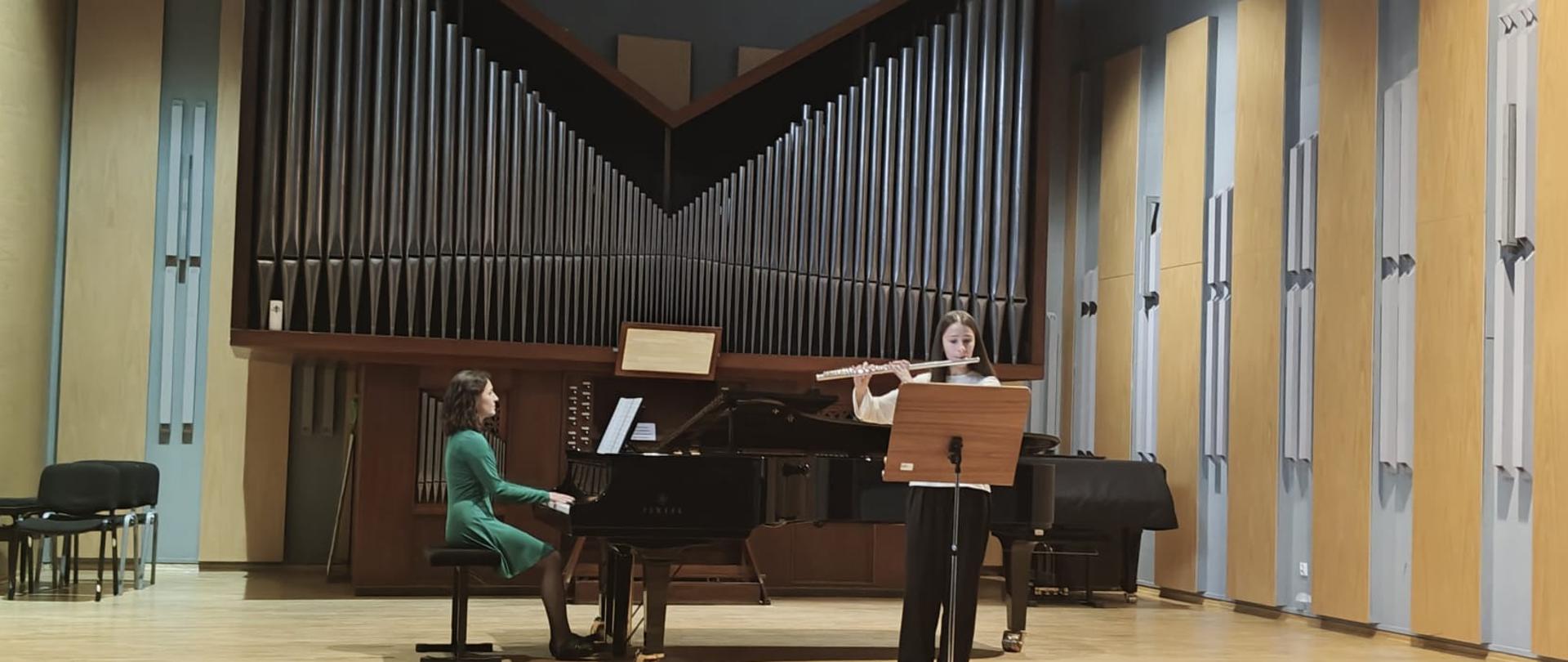 Zdjęcie przedstawia duet muzyczny na scenie podczas występu lub próby w sali koncertowej. Wykonawcy: Po lewej stronie, kobieta w zielonej sukience siedzi przy czarnym fortepianie i gra na nim. Po prawej, młodsza dziewczyna stoi, grając na flecie poprzecznym. Przed flecistką stoi drewniany pulpit z nutami. Otoczenie: Tło stanowi imponujący prospekt organowy z licznymi piszczałkami. Scena ma jasną drewnianą podłogę, a ściany są wyłożone akustycznymi panelami z drewna i jasnego materiału. Po lewej stronie widoczne są puste krzesła dla publiczności. Całość tworzy kameralną i uroczystą atmosferę typową dla recitalu muzyki klasycznej.
