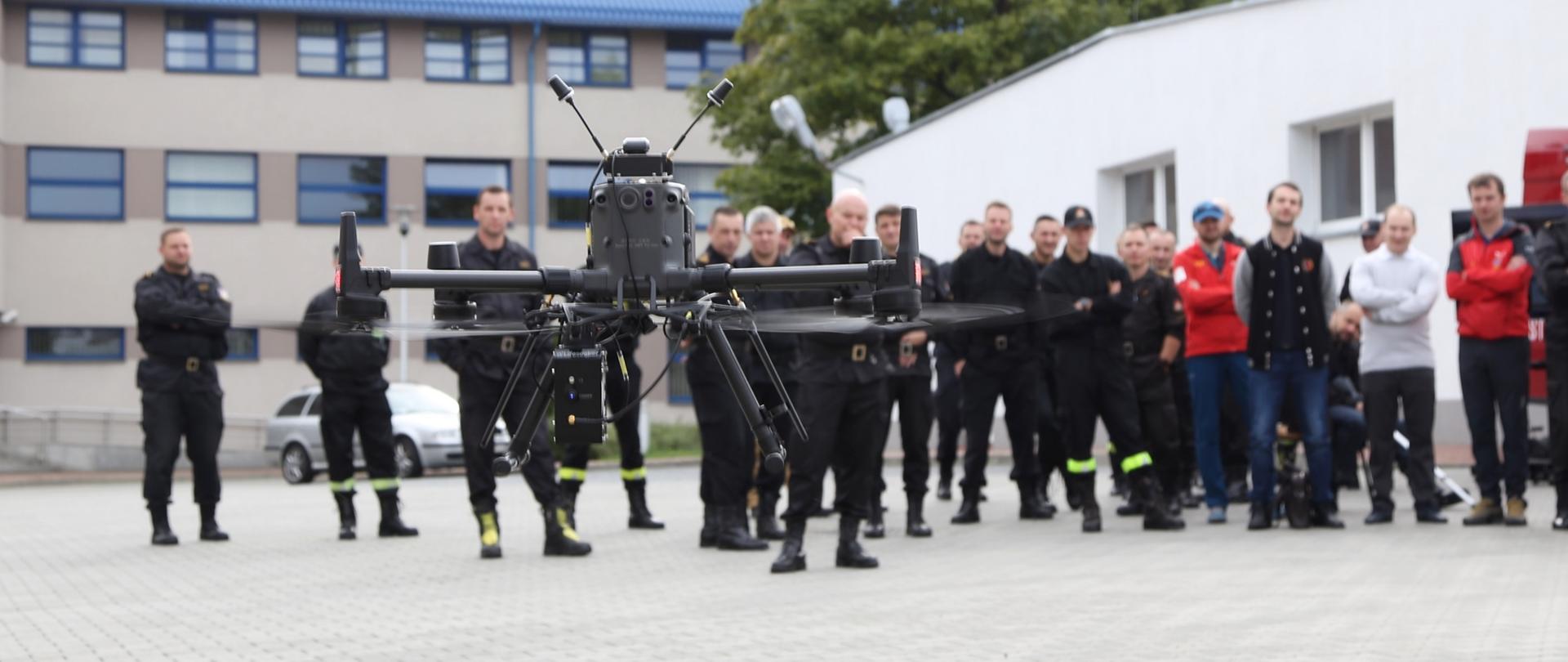 Na zdjęciu dron unoszący się nisko nad powierzchnią ziemi, w tle widoczni uczestnicy warsztatów