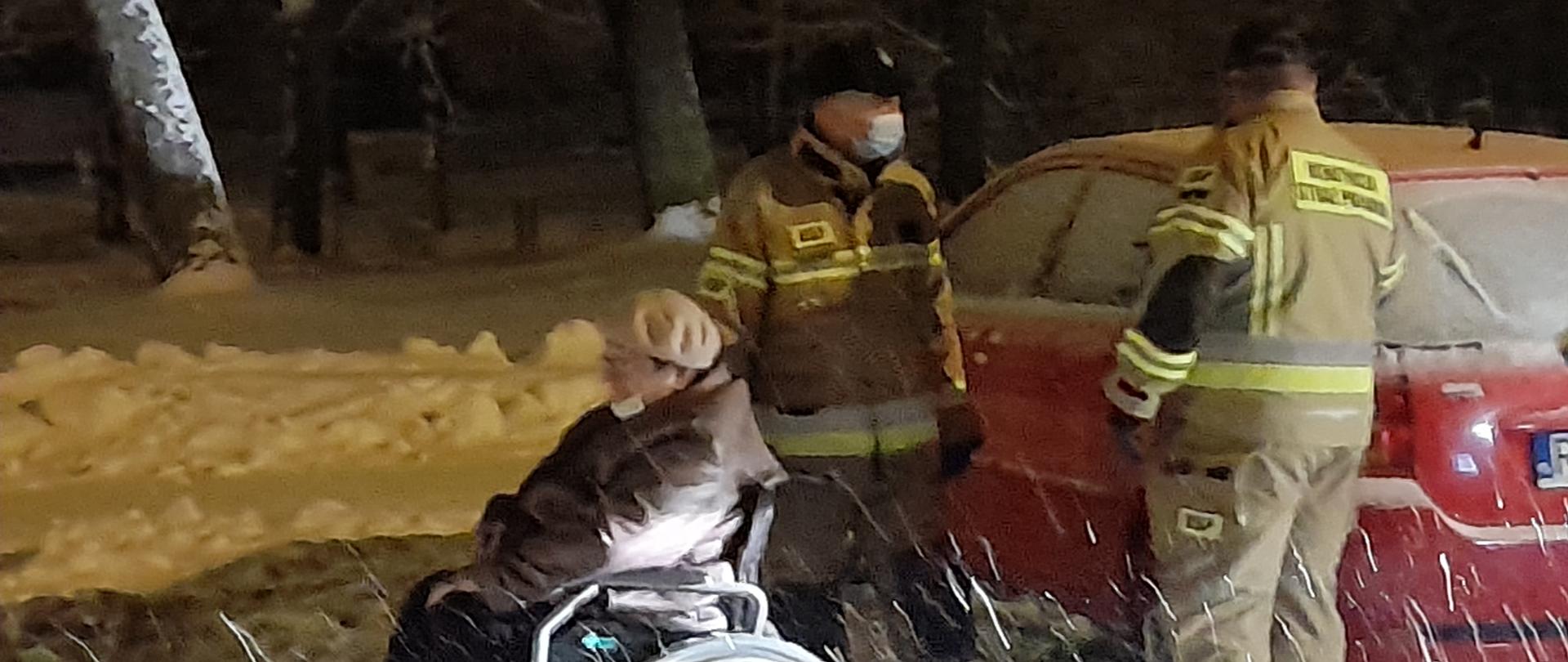 Zdjęcie przedstawia osobę na wózku inwalidzkim oraz 2 strażaków. Zdjęcie wykonane w porze nocnej przy opadach śniegu.