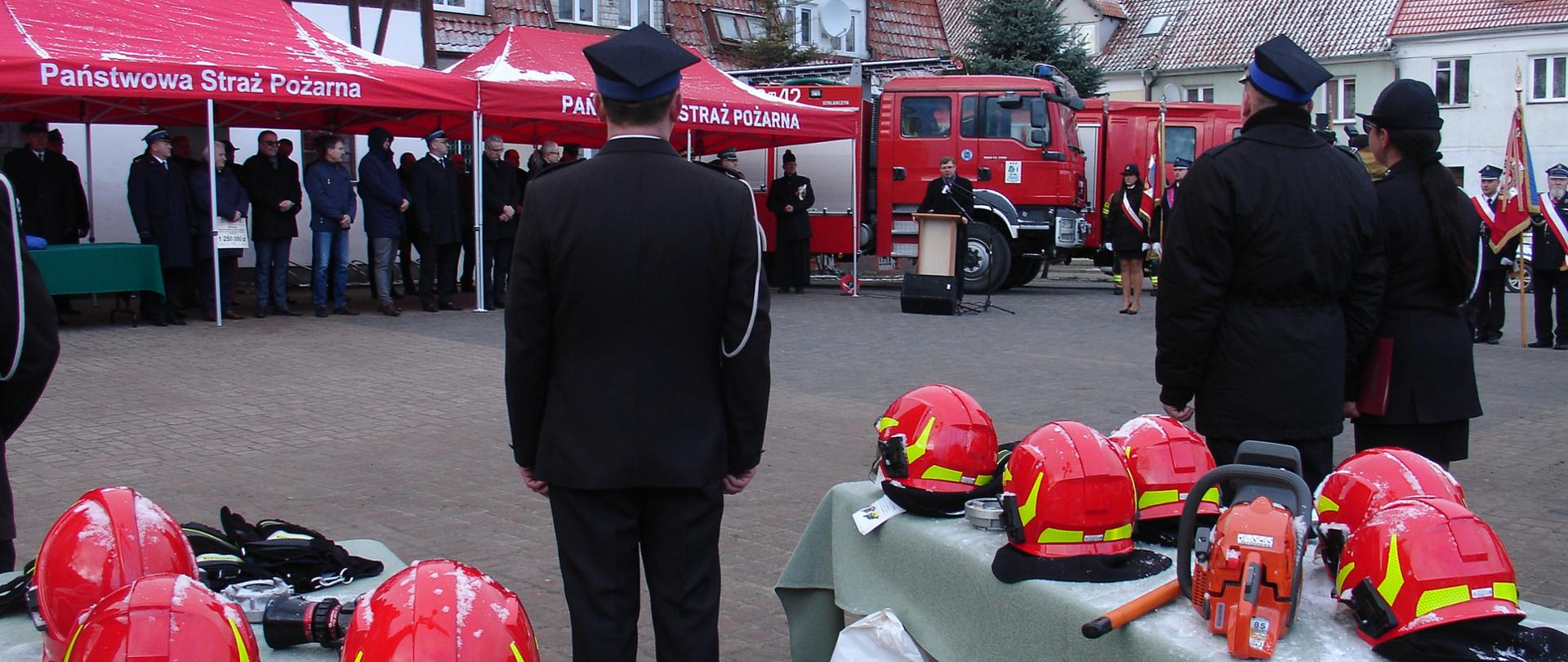 Zdjęcie zrobione na placu przed Urzędem Gminy Srokowo. Funkcjonariusze ubrani w ciemny mundur wyjściowy z rogatywkami na głowie. Na pierwszym planie dwa stoły z wystawionym sprzętem gotowym do przekazania. Obok stołów ustawieni druhowie czekający na przekazanie. W tle czerwone namioty, a pod nimi stoją zaproszeni goście. Obok namiotu dwa czerwone samochody ratowniczo-gaśnicze.