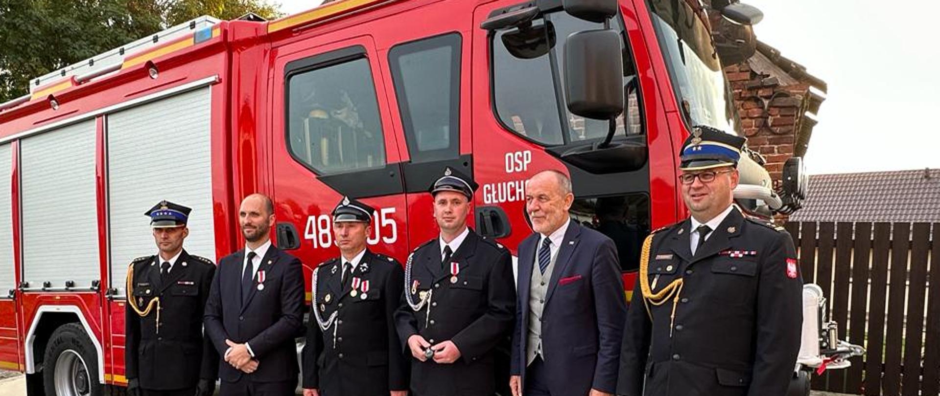 kilku strażaków stoi obok siebie, ubrani w mundury ze sznurem i w rogatywce, za nimi stoi czerwony samochód strażacki