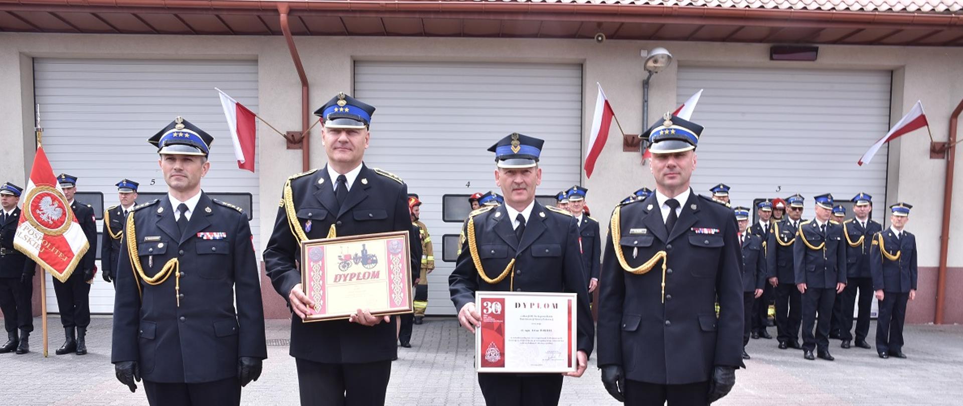 Zdjęcie przedstawia funkcjonariuszy Straży Pożarnej po otrzymaniu wyróżnień i odznaczeń. W tle budynek komendy.