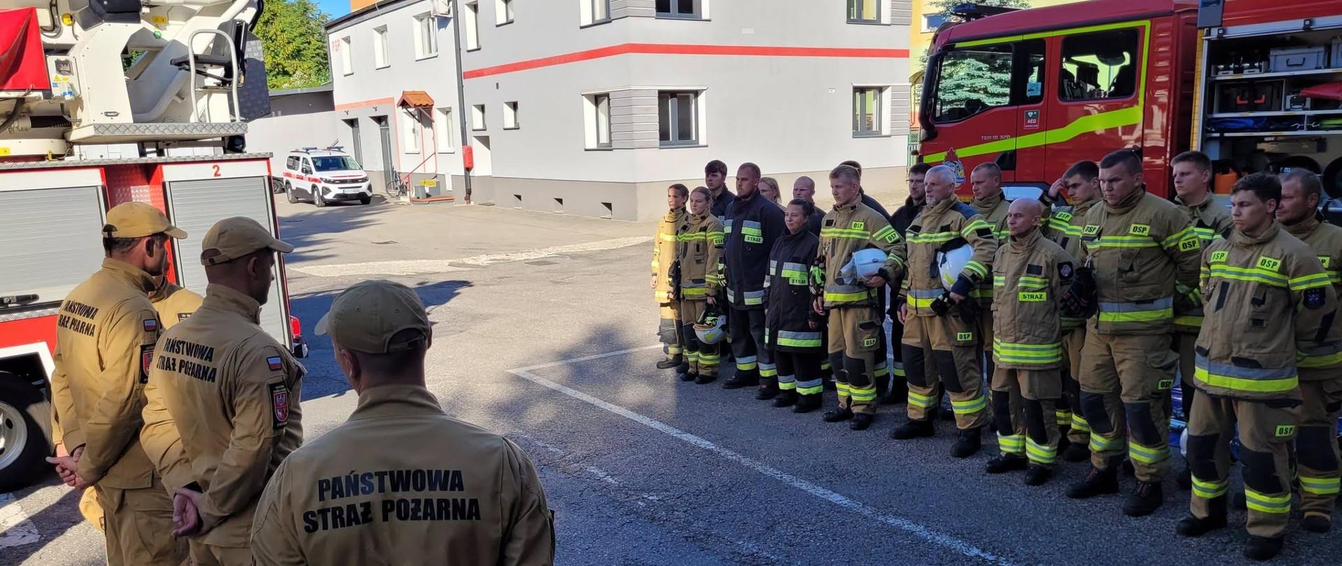 Grupa strażaków stoi w szeregu na placu przed jednostką straży pożarnej. Obok widać czerwone wozy strażackie, a kilku dowódców prowadzi odprawę.
