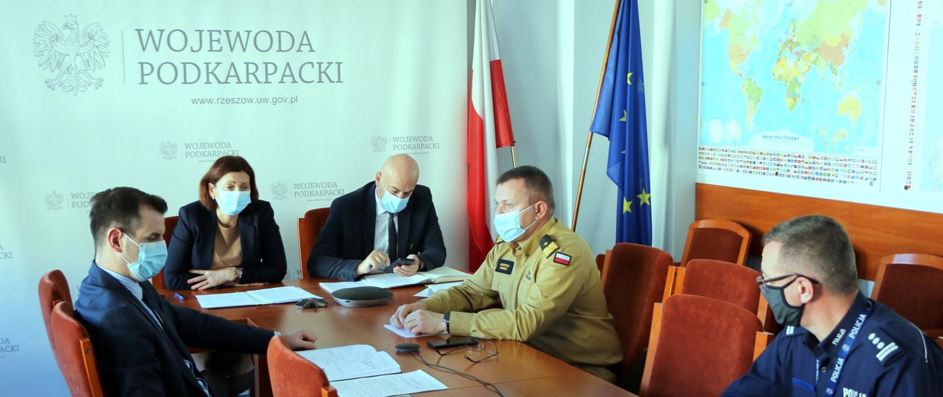 Zdjęcie osób siedzących przy stole konferencyjnym. Przy stole siedzi pięć osób. Jedna z nich w mundurze policyjnym, a druga w mundurze strażaka PSP w stopniu generała. Za uczestnikami spotkania widać banner z napisem wojewoda podkarpacki.