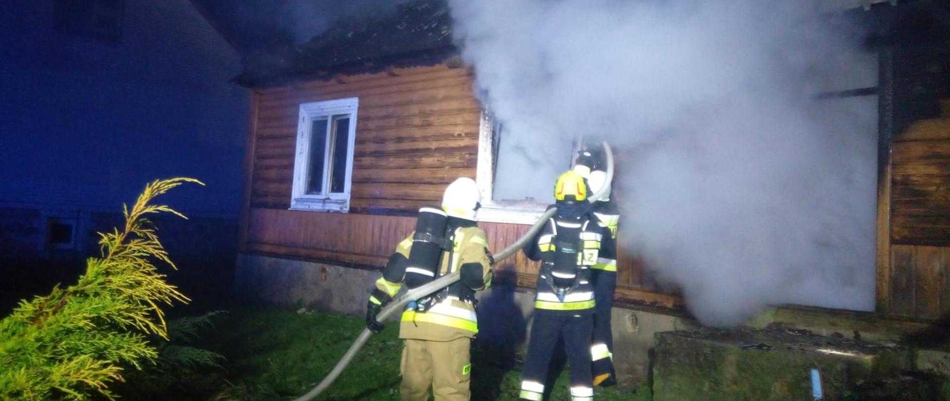 Zdjęcie przedstawia dom drewniany, w którym wybuchł pożar. Rota strażaków podaje wodę przez okno do środka skąd wydobywa się gesty dym i para wodna.