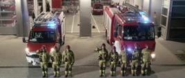 Na zdjęciu strażacy przed jednostką stoją przed samochodami które mają włączone sygnały świetlne i dźwiękowe oddając hołd