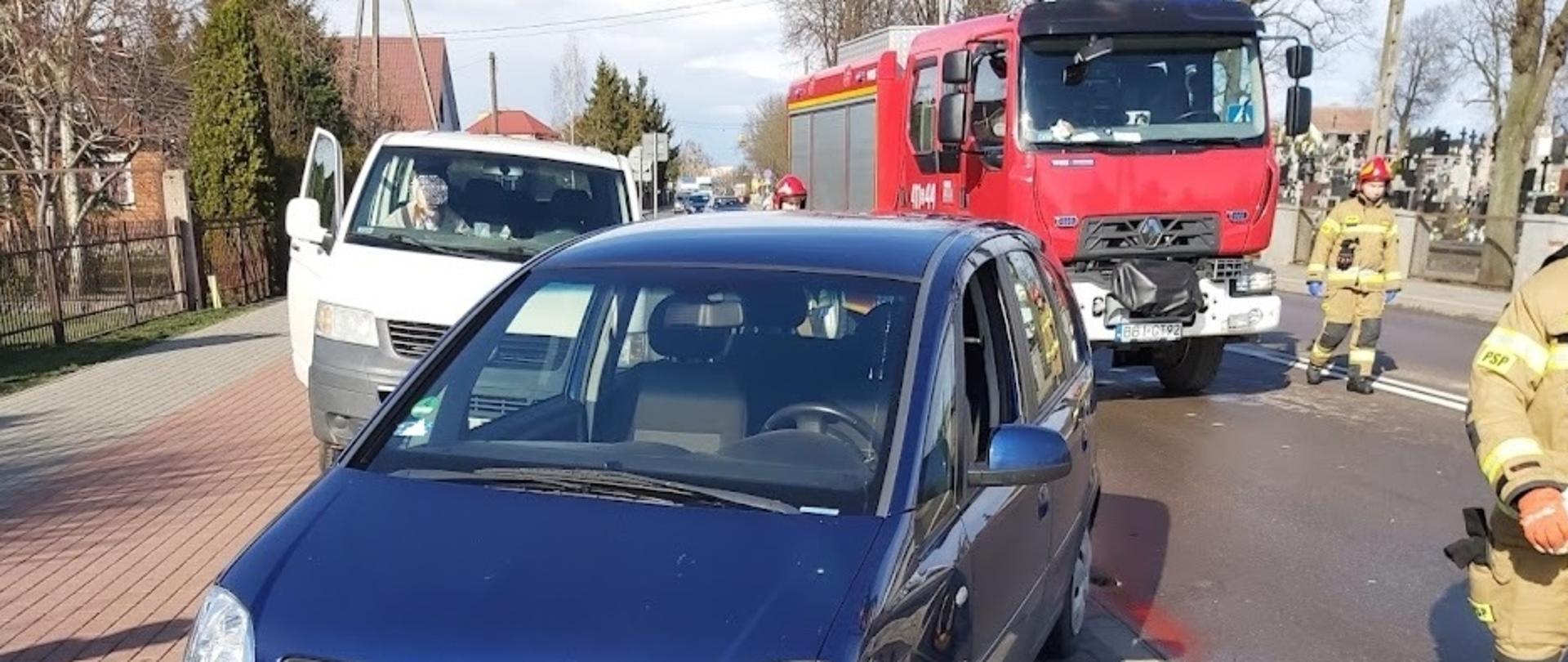 Samochód osobowy marki Opel stojący na zatoczce postojowej po zderzeniu z innym autem. W pojeździe uszkodzony jest prawa przednia część (zderzak, nadkole). W tle samochód ratowniczo-gaśniczy.