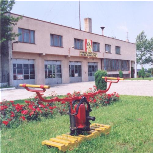 Zdjęcie przedstawia budynek Komendy Rejonowej Straży Pożarnej w Koluszkach wybudowanej w 1975r. przy ul. Słowackiego 28. Na skwerku stara pompa ręczna. 