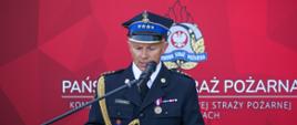
Na zdjęciu przemawia stażak w mundurze wyjściowym koloru czarnego przy pulpicie koloru czerwonego z logo państwowej straży pożarnej na tle namiotu w kolorze czerwoym i siedzących pod namiotem gości.