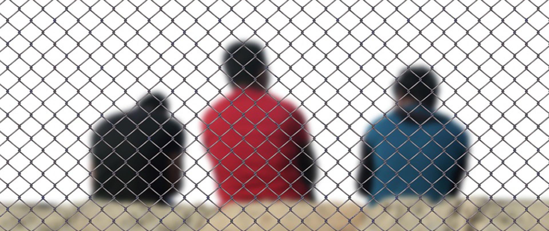 Na zdjęciu: 3 niewyraźne sylwetki osób siedzących tyłem za siatką ogrodzeniową