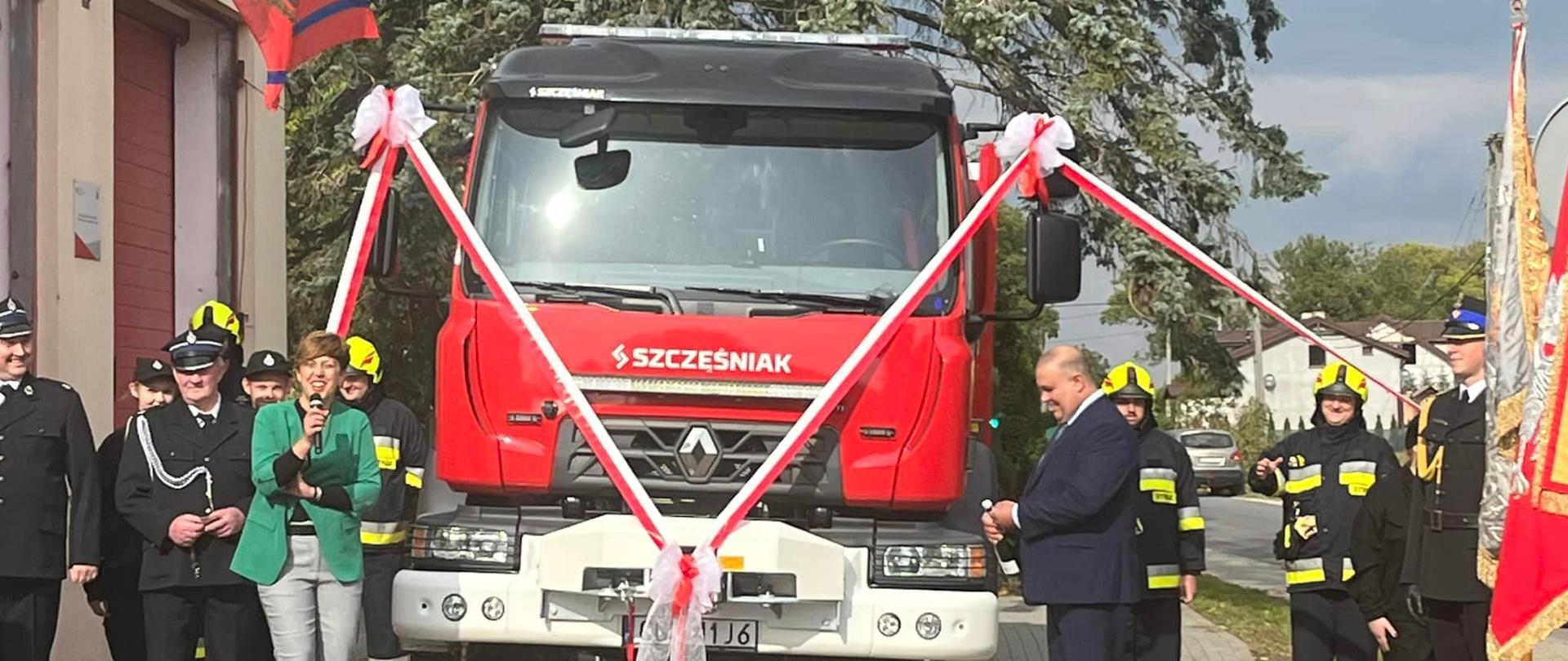 Na zdjęciu widać nowy pojazd ratowniczo-gaśniczy ozdobiony biało-czerwonymi wstęgami. Po lewej stronie pojazdu stoi jego matka chrzestna i trzyma w dłoniach mikrofon , natomiast po lewej stronie stoi mężczyzna ojciec chrzestny z butelką szampana w dłoni.