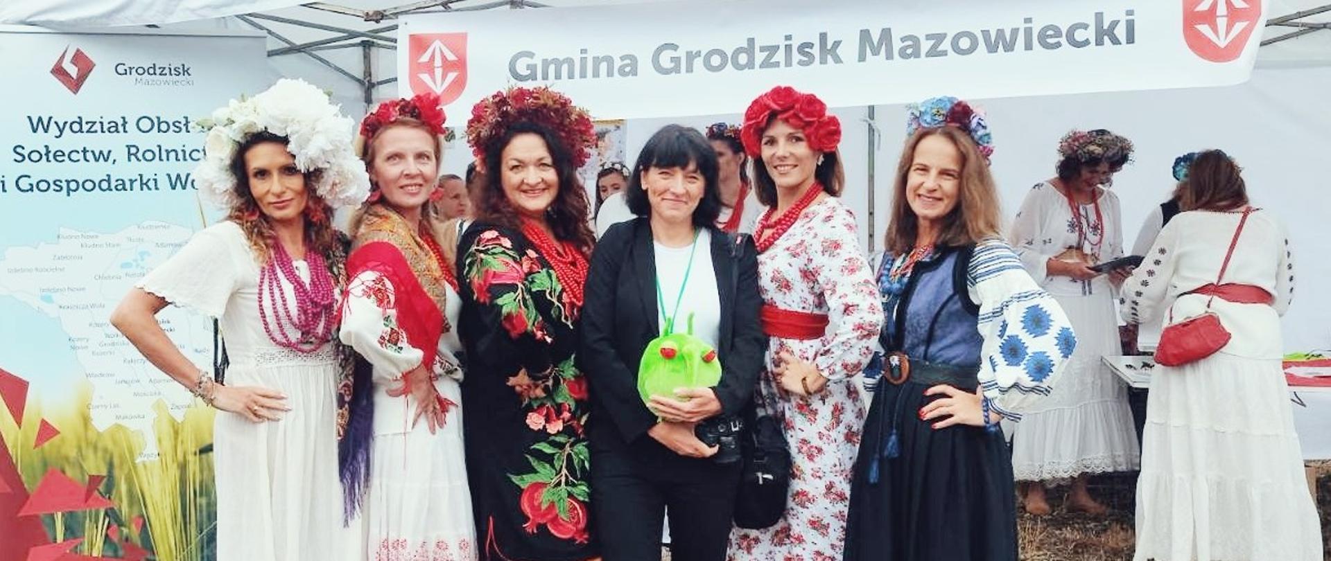 Grupa sześciu kobiet pozuje do zdjęcia przed stoiskiem „Gmina Grodzisk Mazowiecki”, pięć z nich w barwnych, ludowych strojach z kwiatowymi wiankami, a jedna w czarnym ubraniu trzyma zieloną pluszową maskotkę.