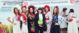 Grupa sześciu kobiet pozuje do zdjęcia przed stoiskiem „Gmina Grodzisk Mazowiecki”, pięć z nich w barwnych, ludowych strojach z kwiatowymi wiankami, a jedna w czarnym ubraniu trzyma zieloną pluszową maskotkę.