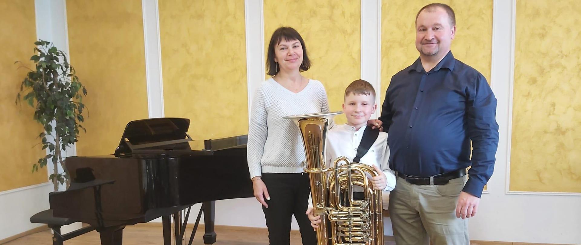 Na zdjęciu stoją 3 osoby, za nimi fortepian, uczeń pośrodku trzyma tubę