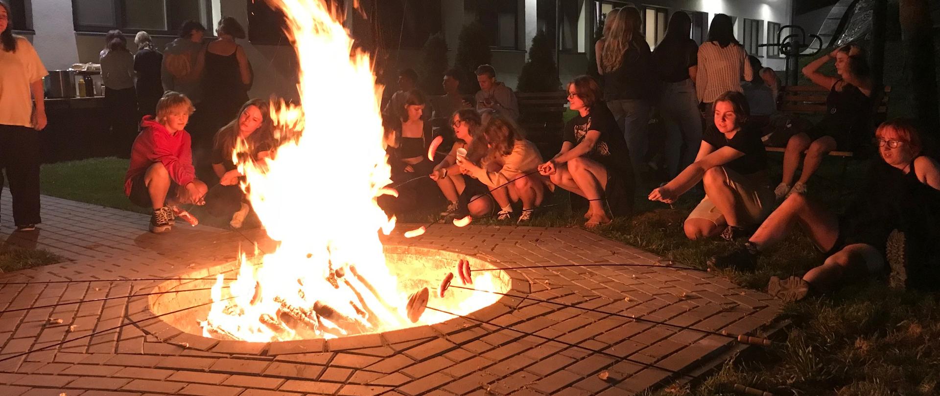 Na pierwszym planie widać płonące ognisko, wokół niego szeroka opaska z kostki brukowej, w dalszym planie młodzi ludzie pieką kiełbaski, siedzą na ławkach lub stoją, w tle widać budynek bursy