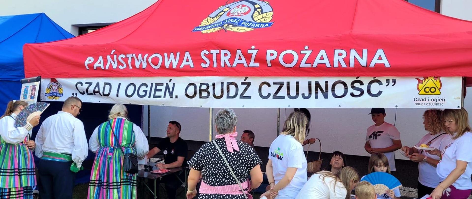 Pod namiotem z napisem na górze: Państwowa Straż Pożarna "Czad i Ogień. Obudź czujność" znajdują się ludzie - osoby dorosłe oraz dzieci, które rozmawiają ze strażakami oraz losują losy w ramach konkursu.