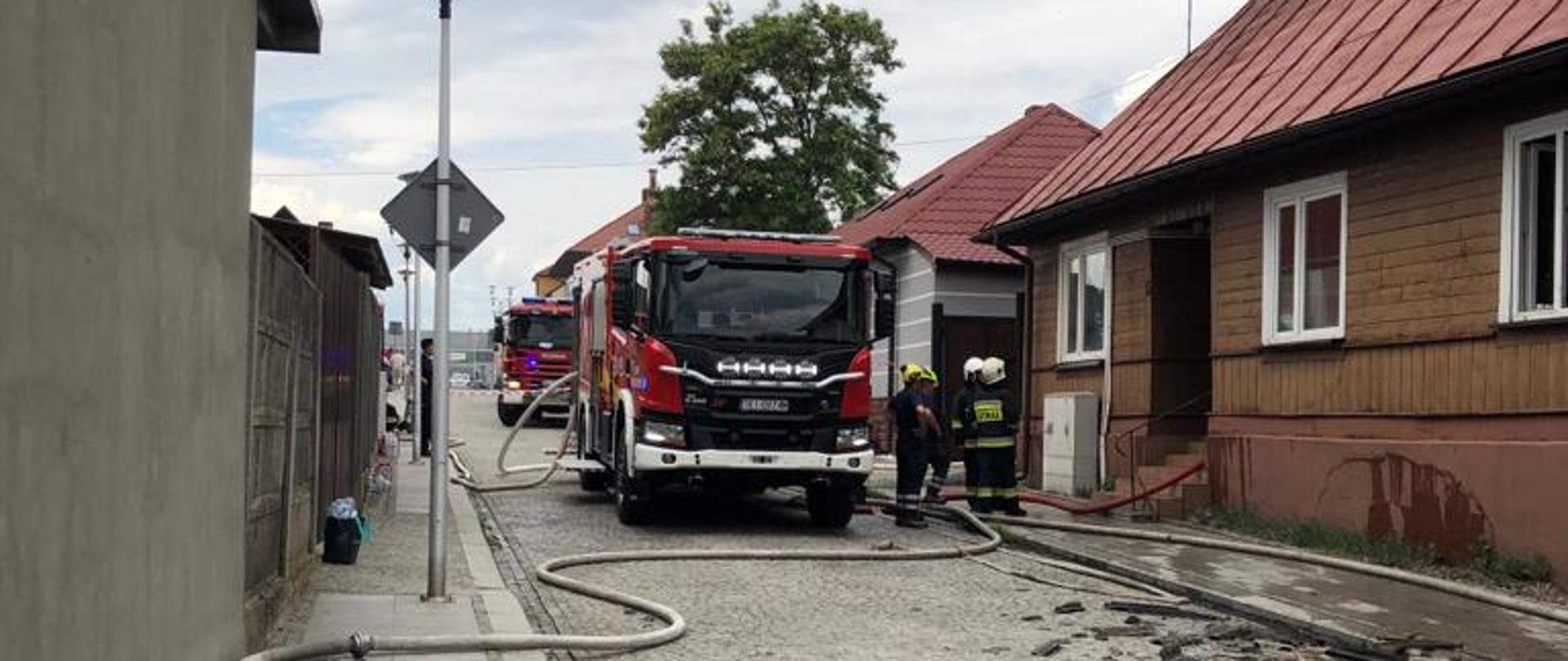 Zdjęcie przedstawia ulicę Mikołaja Reja przy której doszło do pożaru. W wąskiej uliczce zaparkowane stoją dwa samochody pożarnicze, przy nich widać grupę strażaków. Węże gaśnicze leżą rozciągnięte na jezdni.
