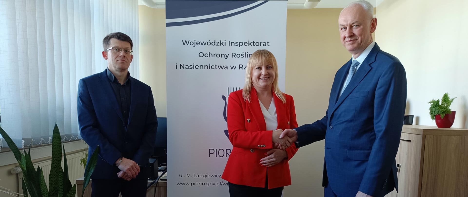 Dwóch mężczyzn i jedna kobieta ubrani na elegancko. Mężczyźni ubrani w granatowe garnitury a kobieta w czarne spodnie, białą koszulkę i czerwoną marynarkę. Kobieta ściska dłoń mężczyźnie po prawej stronie. Za nimi stoi biało-granatowy baner z logiem instytucji. Za banerem stoi biurko a po prawej stronie szafa na której stoi mały kwiatek. W pokoju są jasne kolory. Na pierwszym planie po lewej stronie stoi duży zielony kwiatek.
