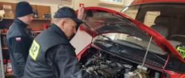 Dwóch strażaków stoi przy otwartej pokrywie silnika w samochodzie OSP.