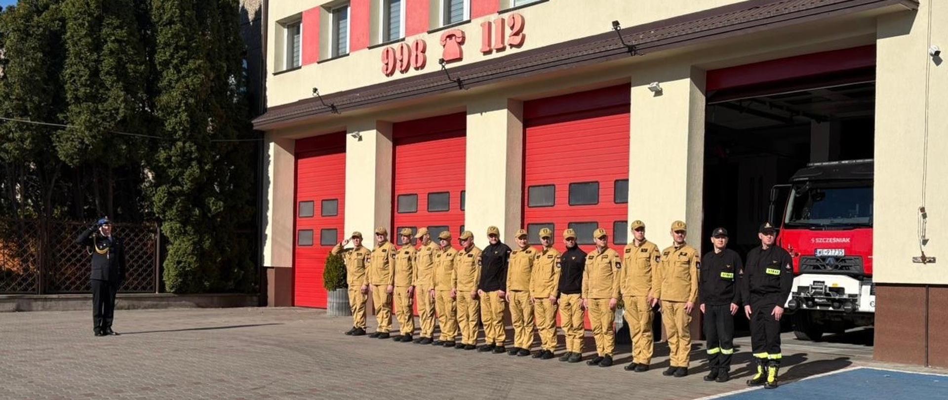 Strażacy ustawieni w szeregu przed budynkiem Komendy Powiatowej Państwowej Straży Pożarnej w Mławie podczas uroczystości, w tle czerwone bramy garażowe i samochód pożarniczy.