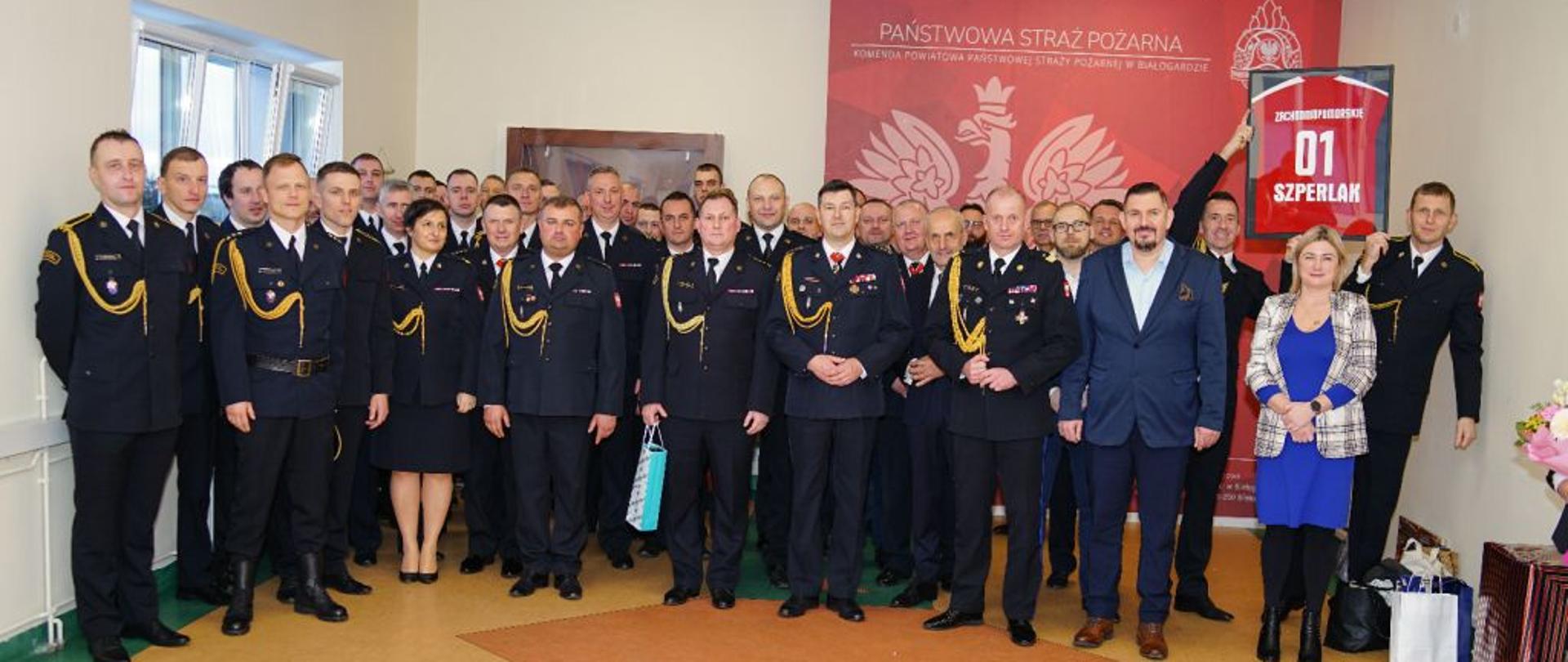 Zdjęcie grupowe. Na tle ścianki z godłem Polski stoją uczestnicy uroczystości. Strażacy w mundurach wyjściowych i koszarowych, cywile, rodzina. Wszyscy szczęśliwi i uśmiechnięci.