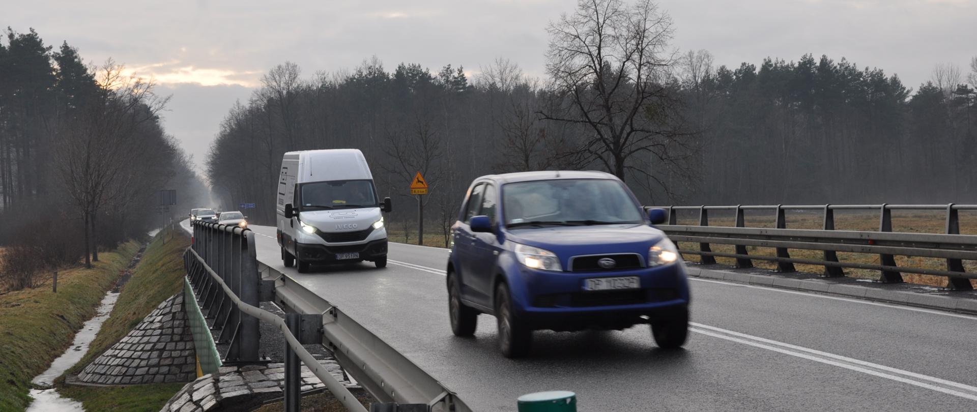 Zdjęcie przedstawia fragment drogi krajowej DK45 na odcinku Zawada - Jełowa. Na fotografii widoczny jest mostek nad rowem melioracyjnym z barierkami ochronnymi po obu stronach. Na drodze znajdują się dwa pojazdy: niebieski samochód osobowy oraz biały samochód dostawczy. Niebo jest zachmurzone, co sugeruje pochmurną pogodę.