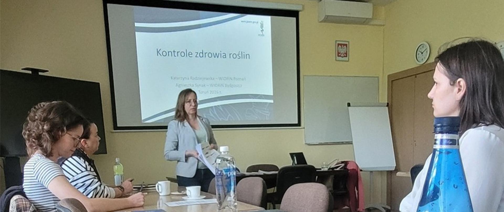 Fotografia przedstawia prowadzącą zajęcia Panią Katarzynę Radziejewską, reprezentującą Dział Nadzoru Fitosanitarnego Wojewódzkiego Inspektoratu Ochrony Roślin i Nasiennictwa w Poznaniu. Prelegentka stoi na tle wyświetlanej planszy prezentacji, na której widnieje temat: ,,Kontrole zdrowia roślin”. Ubrana jest w jasny żakiet. Na pierwszym planie widzimy 3 uczestniczki szkolenia, którzy siedzą przy długim stole, słuchają wystąpienia i sporządzają notatki
