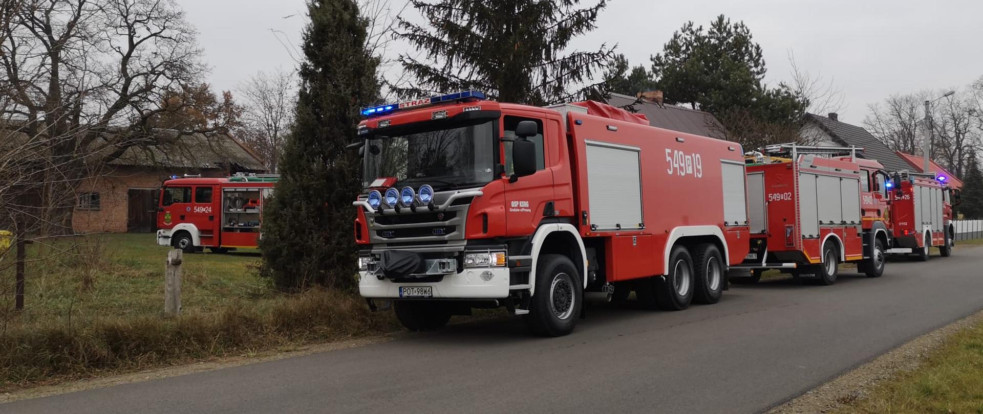 Na pierwszym planie samochody strażackie stoją na jezdni przy posesji, na której miał miejsce pożar. Na drugim planie widać wóz strażacki, który stoi wewnątrz posesji. Dookoła zabudowania, na posesji rosną drzewa.