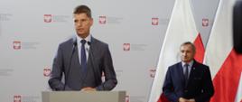Minister Edukacji Narodowej Dariusz Piontkowski i wiceminister Maciej Kopeć podczas konferencji. W tle biała ścianka z logo ministerstwa oraz flagi Polski.