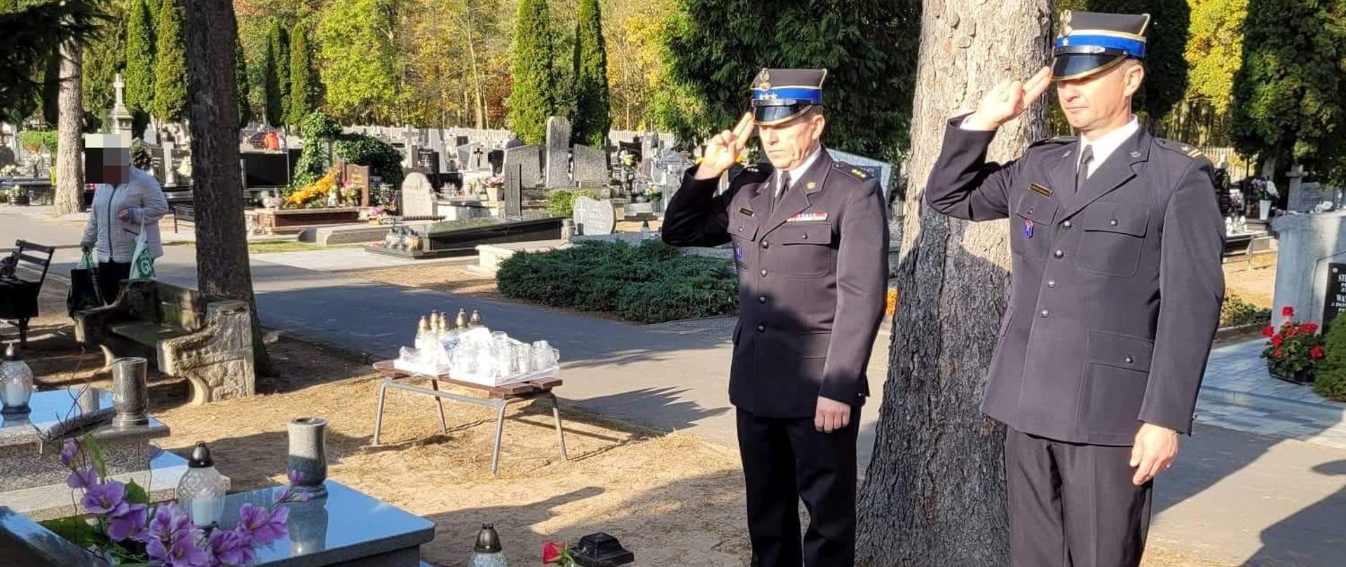 Dwóch strażaków w mundurach oddaje hołd przy grobie na cmentarzu