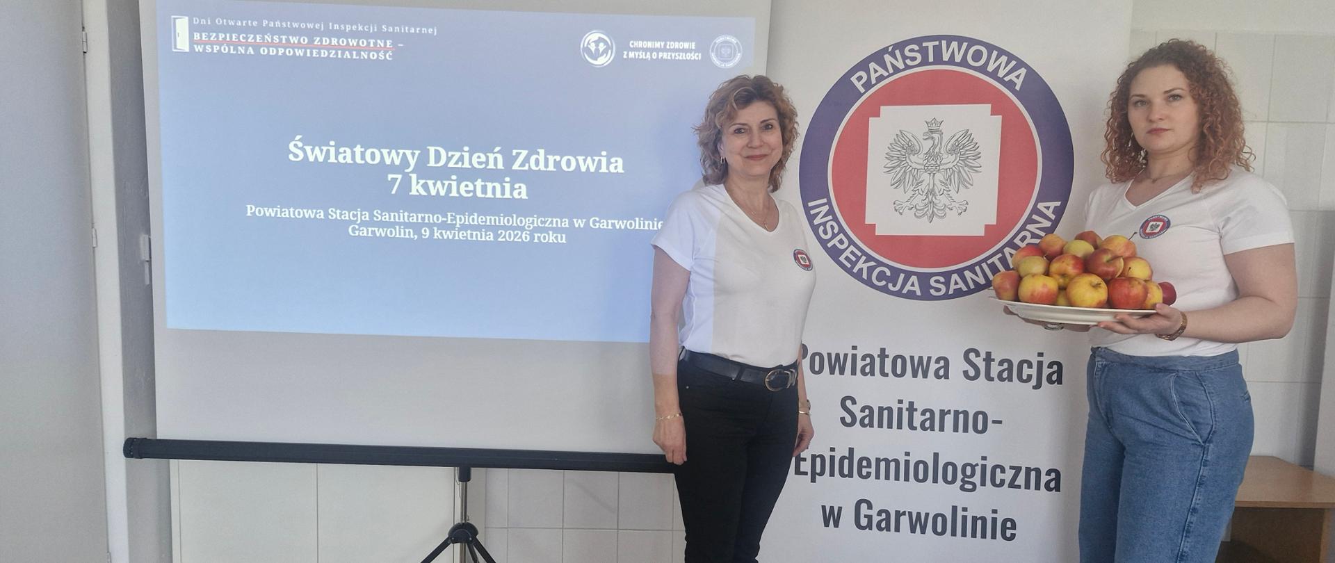 Dwie pracownice PSSE na tle slajdu "Światowy Dzień Zdrowia" i baneru z logo Państwowej Inspekcji Sanitarnej. Jedna z osób trzyma talerz z jabłkami.