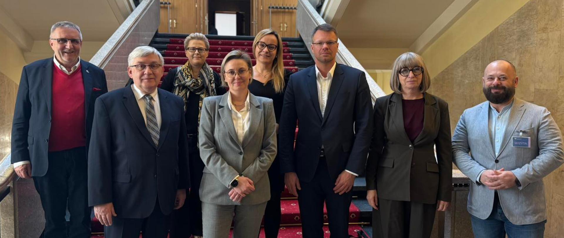 Szeroki kadr, zdjęcie grupowe. Uczestnicy spotkania pozują do wspólnego zdjęcia. Widoczna między innymi Sekretarz Stanu Adriana Porowska. W tle widać wnętrze budynku Kancelarii Prezesa Rady Ministrów. 