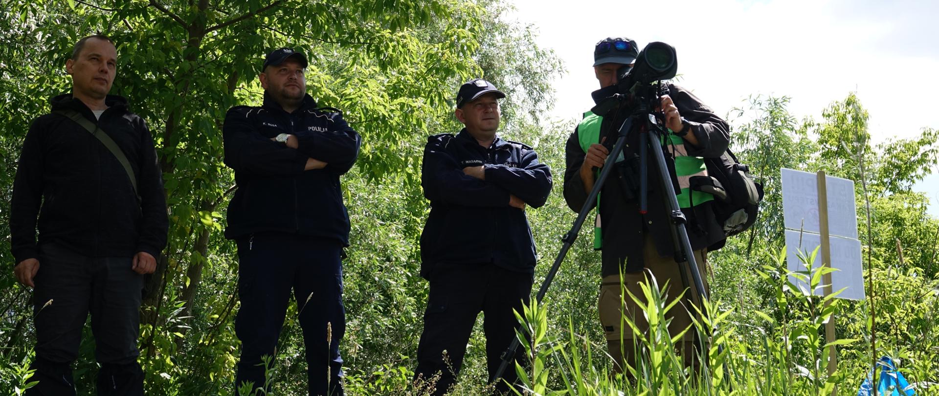 4 mężczyzn stojących na skarpie, jeden w ubraniu terenowym z lunetą, dwóch policjantów, w tle drzewa.