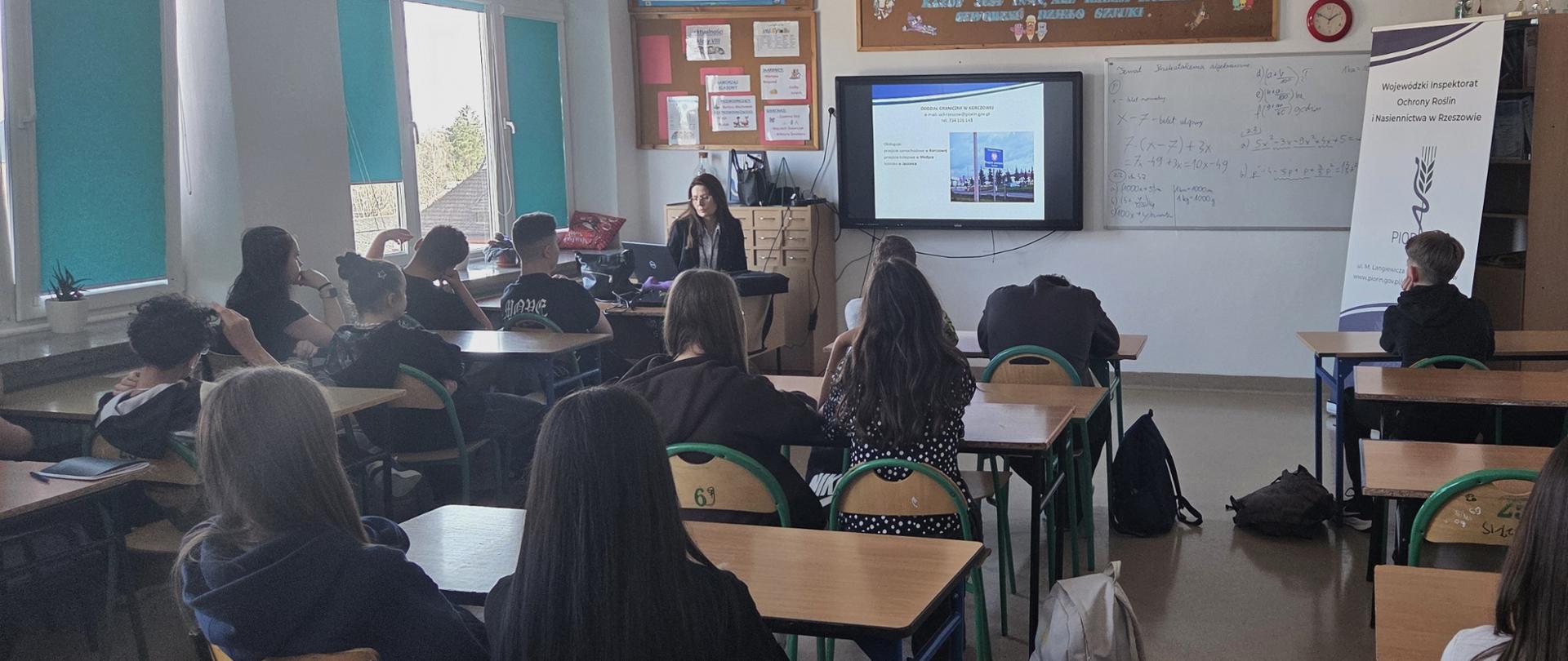 Szerokie ujęcie klasy szkolnej z wieloma uczniami siedzącymi w rzędach przy ławkach. Nauczycielka siedzi przy biurku na przodzie sali, prowadząc prezentację wyświetlaną na ekranie. Po lewej stronie znajdują się duże okna z zielonymi roletami, przez które wpada naturalne światło. Na ścianach wiszą tablice edukacyjne, dekoracje oraz krzyż i godło Polski. Uczniowie są zwróceni w stronę nauczycielki i ekranu, niektórzy opierają głowy na rękach, słuchając wykładu. W klasie widoczne są plecaki ustawione obok ławek oraz schludnie rozmieszczone meble szkolne.